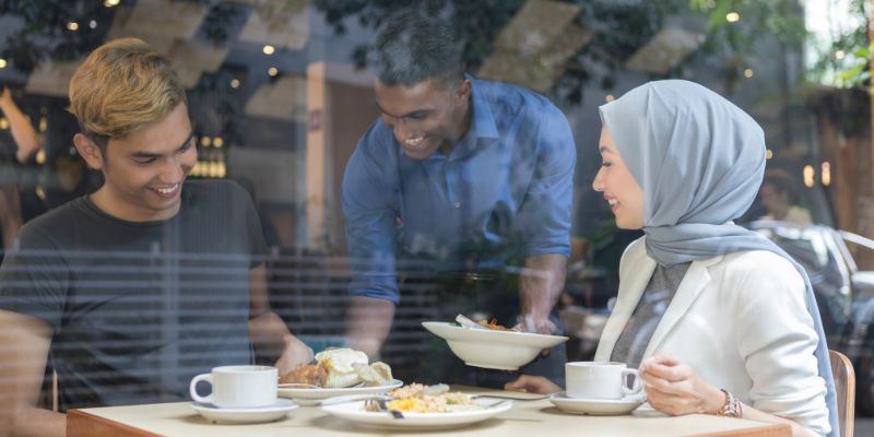 Pelayan lelaki berbaju biru sedang menghidangkan makanan kepada pelanggan lelaki dan perempuan di restoran mereka. 