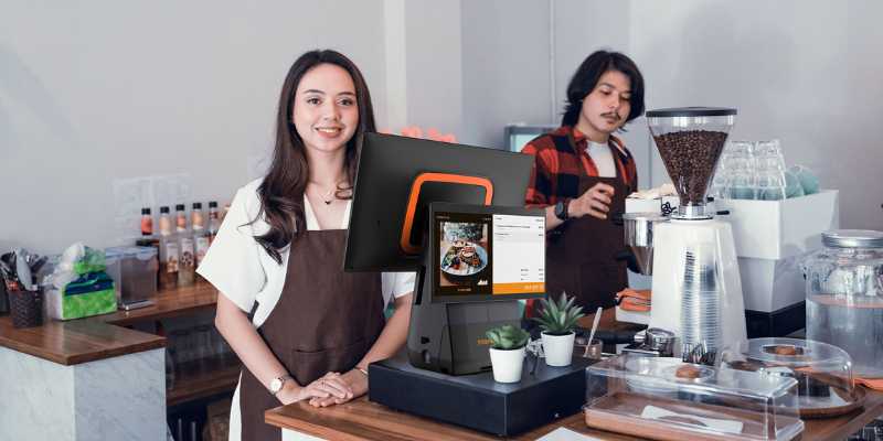 A girl is standing in front of the POS system while a barista is making coffee beside her.