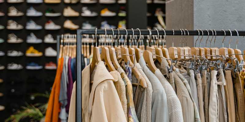 The picture showcases clothes hanging on a railing with beautifully displayed shoes in the background at the retail store.