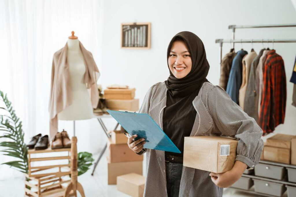 A woman store manager wearing a hijab is smiling at the camera while holding stocks and a checklist.