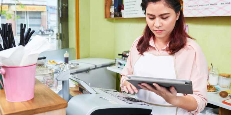 Seorang juruwang wanita sedang berdiri di hadapan sistem daftar tunai manual sambil memegang ipad.