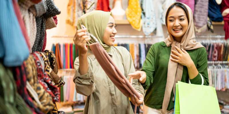 Dua orang pembeli wanita sedang memilih tudung di kedai tudung.
