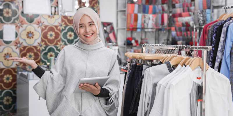 Seorang wanita bertudung tersenyum sambil memegang ipad di butik pakaian.