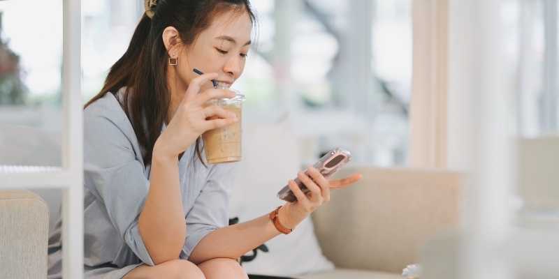 Asian girl smiling while holding an iced coffee in a takeaway cup and a phone in her other hand