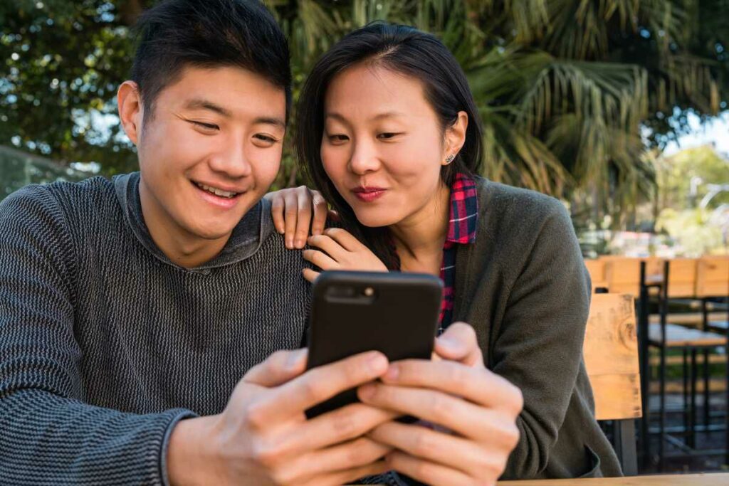 Asian couple smiling happily while reading a text message at a restaurant.