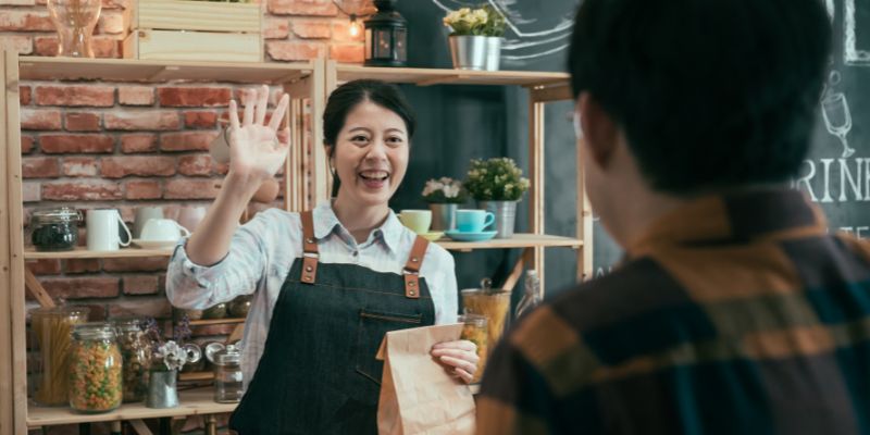 Seorang pelayan F&B wanita sedang melambai kepada pelanggan sambil memegang beg makanan pesanan pelanggan.