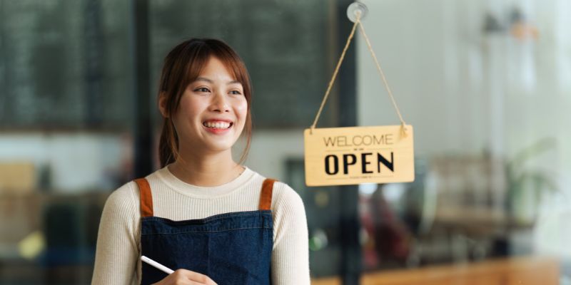 Seorang pelayan F&B perempuan sedang berdiri di hadapan kafe sambil tersenyum.